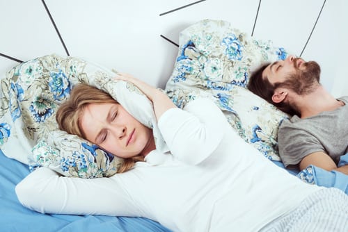 A man and a woman in bed, man is snoring and woman is covering her ears.